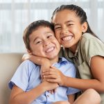 Cheerful Asian girl smiling and embracing cute boy while sitting on comfortable couch together