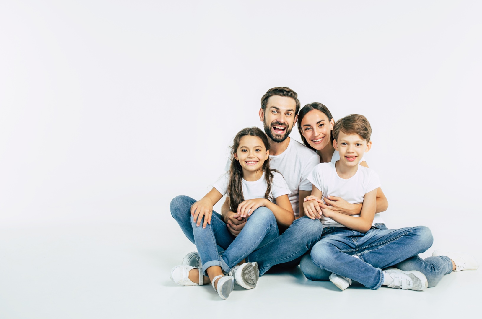 Relationship concept. Beautiful and happy smiling young family in white T-shirts are hugging and have a fun time together while sitting on the floor and looking on camera.