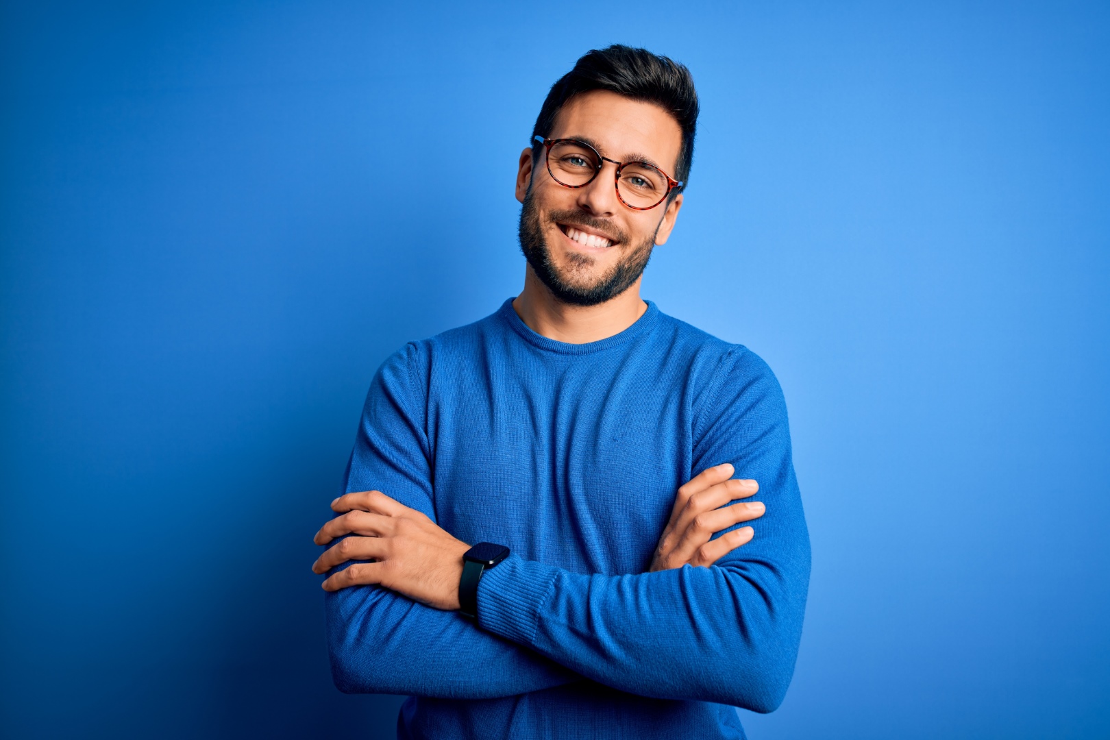 Young handsome man with beard wearing casual sweater and glasses over blue background happy face smiling with crossed arms looking at the camera. Positive person.